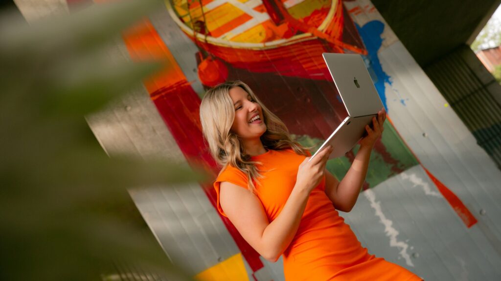 Katherine wears an orange dress and smiles at her laptop, no doubt reading  her article on blogging and Pinterest for wedding pros